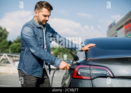 Vista laterale di un giovane bell'uomo in camicia jeans, collegando il filo alla presa per caricare la sua nuova auto elettrica moderna di lusso all'aperto Foto Stock