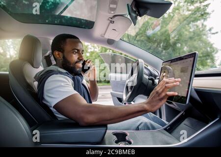 Bell'uomo africano con bearded tocca il touchscreen nel suo nuovo veicolo elettrico high-tech mentre parla al telefono e sorride. Guida automatica Foto Stock
