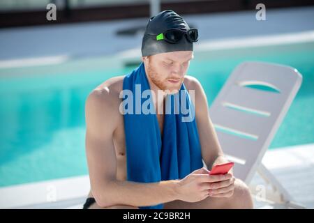 Nuotatore maschile attento con smartphone seduto a bordo piscina Foto Stock