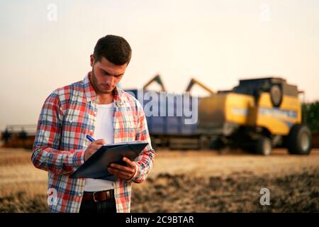 L'agricoltore controlla il caricamento del frumento dalla trebbiatrice al dumper per granella. Conducente che tiene appunti, tiene appunti, conta carichi. Lo spedizioniere compila la spedizione Foto Stock