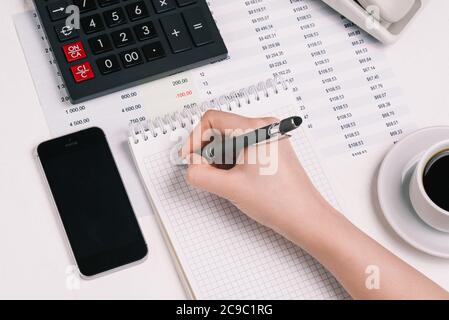 La mano della donna scrive in un taccuino con una matita. Desktop con calcolatrice, telefono cellulare, Coffee Cup e documenti finanziari. Concetto di contabilità Foto Stock