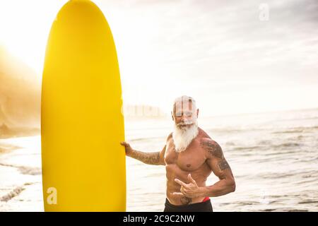 Tattooed surfer senior tenendo surf board sulla spiaggia al tramonto - ragazzo felice vecchio divertirsi facendo sport estremo - gioioso anziano concetto - Focus on hi Foto Stock