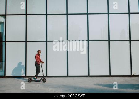 Un bel giovane uomo d'affari con la bearded guida e scooter di fronte della parete bianca dell'edificio Foto Stock