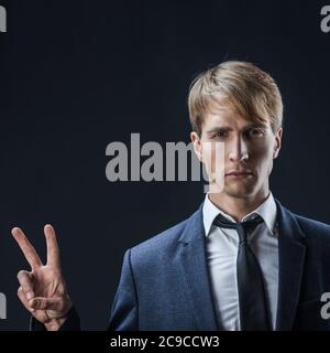 Uomo d'affari felice mostra un gesto di due-pollice-su. Nel commercio, tutto è super, un giovane in un vestito con una cravatta su uno sfondo scuro. Foto Stock