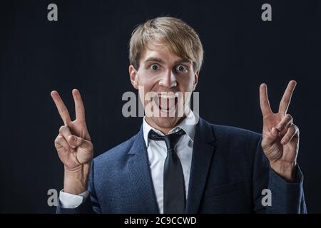 Uomo d'affari felice mostra un gesto di due-pollice-su. Nel commercio, tutto è super, un giovane in un vestito con una cravatta su uno sfondo scuro. Foto Stock