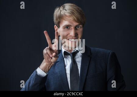 Uomo d'affari felice mostra un gesto di due-pollice-su. Nel commercio, tutto è super, un giovane in un vestito con una cravatta su uno sfondo scuro. Foto Stock