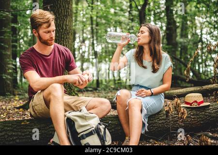 La coppia caucasica si fermò a riposare mentre camminava nella foresta, i giovani si siedono su un tronco d'albero caduto bevendo acqua da una bottiglia. Riposo attivo, escursione Foto Stock