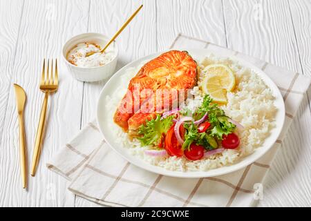 bistecca di pesce rosso fritto servita con riso e insalata di verdure su un piatto bianco su un tavolo di legno con salsa di yogurt senape e posate dorate, paesaggio v Foto Stock