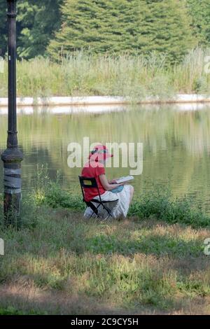 Una donna anziana si siede e legge un libro vicino al lago in Kissena Park, Flushing, New York City. Foto Stock