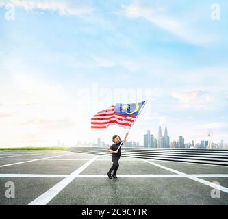 Happy boy che corre mentre tiene la bandiera della Malesia nello spazio aperto con il paesaggio urbano di Kuala Lumpur. Foto Stock