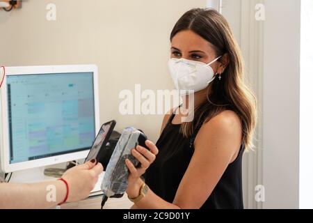 Giovane assistente del negozio con una maschera facciale utilizzando un terminale con carta di credito per pagare con lo smartphone senza contatto. Terminale scheda protetto per una migliore igiene w Foto Stock