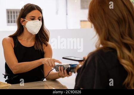 Giovane assistente del negozio con una maschera facciale utilizzando un terminale con carta di credito per pagare con lo smartphone senza contatto. Terminale scheda protetto per una migliore igiene w Foto Stock