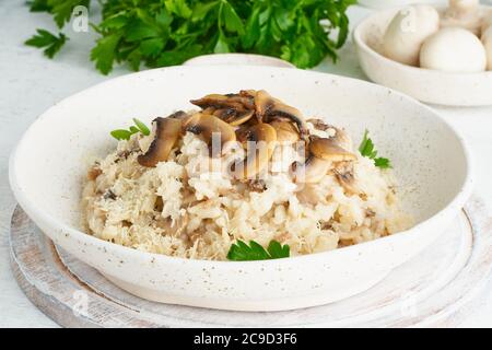 Risotto con funghi in piatto. Porridge di riso con funghi e prezzemolo. Tavola bianca, cucchiai, funghi. Piatti caldi, cucina italiana, vista laterale Foto Stock
