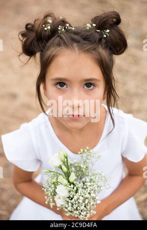 una bambina con i volti seri tiene un bouquet di fiori bianchi .capelli scuri, fiori bianchi nelle mani, vestito bianco Foto Stock