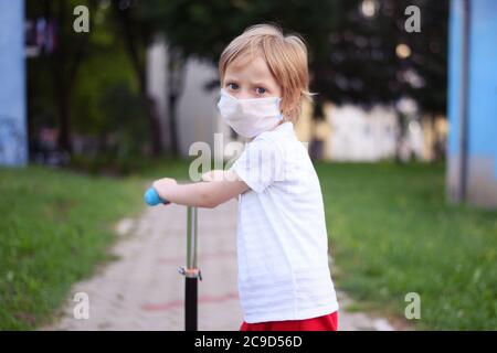 Piccolo ragazzo carino con maschera protettiva che guida uno scooter in un parco. Foto Stock