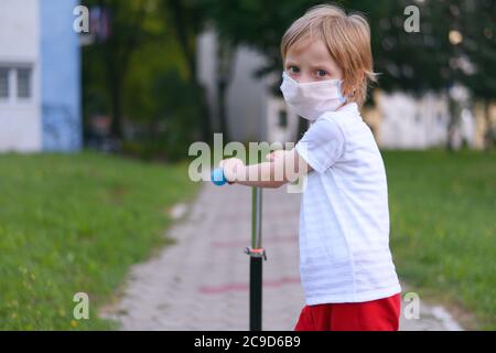 Piccolo ragazzo carino con maschera protettiva che guida uno scooter in un parco. Foto Stock