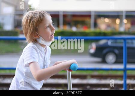 Piccolo ragazzo carino con maschera protettiva che guida uno scooter in un parco. Foto Stock