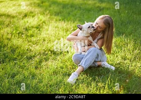 Bel bulldog francese baciare attraente amico umano femmina. Dall'alto si può ammirare la splendida donna caucasica che gode del tramonto estivo, abbracciando un cane carino nel parco cittadino. Amicizia umana e animale. Foto Stock