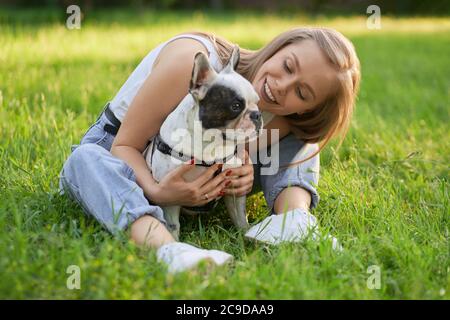 Primo piano vista di una giovane donna che abbrancava un bulldog francese adulto nel parco estivo, seduto sull'erba. Bionda donna cane proprietario in posa con bello bianco e marrone purebred animale domestico che indossa guinzaglio nero, guardando da parte. Foto Stock