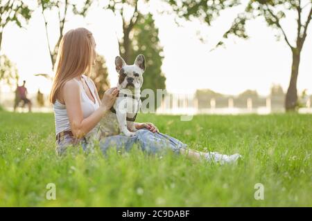 Vista laterale di una giovane donna irriconoscibile seduta sull'erba con un bel bulldog francese sulle gambe. Caucasica ragazza che guarda il tramonto estivo, cane da accarezzare nel parco cittadino. Amicizia umana e animale. Foto Stock