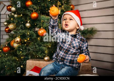 Ritratto di Natale del bambino in cappello rosso di santa seduta su scatole con regali con arance in mani vicino all'albero di natale. Vacanze invernali Natale Foto Stock