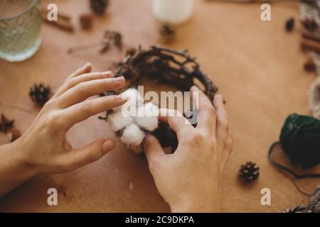 Fiorista che fa il portacandele di natale rustico. Mani che tengono il cotone e fanno la decorazione festiva rustica su tavola di legno con rami e coni di pino. Foto Stock