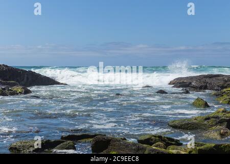 Rocce verdi mussose che circondano una Rockpool con le onde dell'oceano color acqua che si infrangono su grandi rocce con il cielo blu con le nuvole sullo sfondo Foto Stock