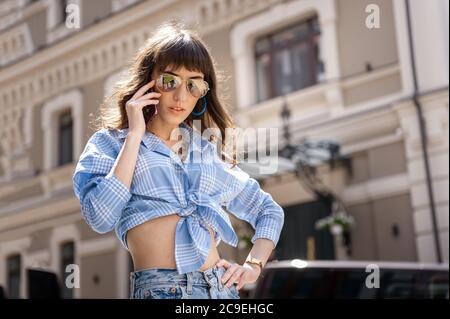 Ritratto esterno di yong bella donna sorridente felice che indossa occhiali da sole eleganti, blusa nera a pois, jeans blu mamma, con piccola borsa trapuntata. Mod Foto Stock