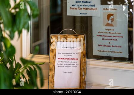 Un cesto di vimini davanti a una finestra dell’Hotel Mühle a Binzen, Markgräflerland, Baden-Württemberg, Germania, funge da punto di consegna per gli ordini scritti della colazione degli ospiti durante la pandemia di coronavirus del 2020. Cartelli accanto richiedono una distanza minima di 1,5 metri e l’uso di mascherine nel ristorante. Foto Stock