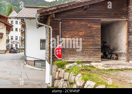 Un segnale di stop rosso è solo parzialmente visibile all'angolo di un edificio in legno scuro nel villaggio di montagna di Berwang, Tirolo, Austria. Una bicicletta è posizionata in un capannone aperto dietro di esso, con una casa in pietra e auto parcheggiate sullo sfondo. Il villaggio si trova a un'altitudine di 1.336 metri nella Tiroler Zugspitz Arena. Foto Stock
