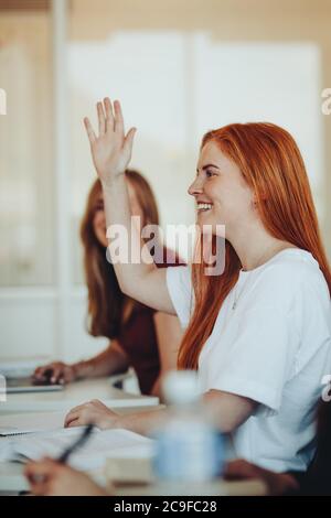 Sorridente studentessa seduta in classe e alzando la mano per porre domande durante la lezione. Lo studente della scuola superiore alza la mano e chiede al docente un qu Foto Stock