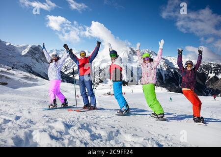 Gruppo di giovani adulti su sci e snowboard si trovano in fila sollevando le mani sulle cime delle montagne Foto Stock