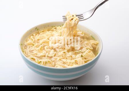 Alimenti a base di sodio elevato, spuntini veloci e pasti in pochi minuti con forchetta in metallo che trattiene tagliatelle istantanee in una ciotola in ceramica isolata su sfondo bianco Foto Stock