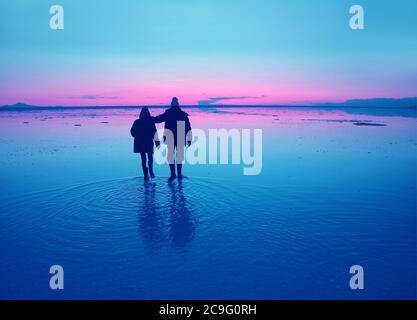 La silhouette in stile pop art di una coppia goditi una passeggiata sulla superficie alluvionale delle distese saline di Uyuni al tramonto, in Bolivia, Sud America Foto Stock