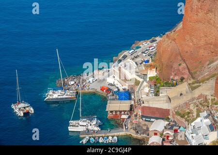 Grecia. Piccolo porto sulla costa rocciosa estiva di Santorini. Catamarani a vela e barche a motore in acqua. Molte persone e auto sulla riva Foto Stock