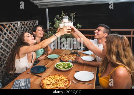 Giovani e felici amici tostare birra e Laughing mentre si mangia pizza all'aperto sul balcone. Summer Night Concept con amici che hanno divertimento, birra e. Foto Stock