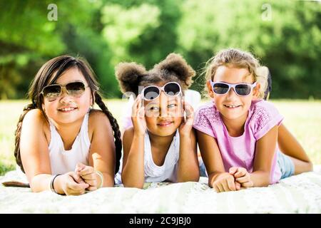 Ragazze che indossano occhiali da sole e che si trovano fianco a fianco sulla coperta nel parco, sorridente, ritratto. Foto Stock