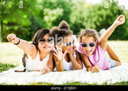 Ragazze che indossano occhiali da sole e che si trovano fianco a fianco sulla coperta nel parco, sorridente, ritratto. Foto Stock