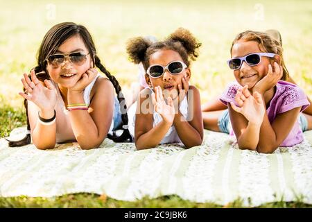 Ragazze che indossano occhiali da sole e che si trovano fianco a fianco sulla coperta nel parco, sorridente, ritratto. Foto Stock