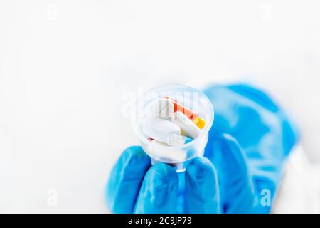 La mano del medico femminile in guanto blu che tiene i bicchieri graduati della medicina pieni di pillole. Copia spazio e sfondo bianco. Foto Stock