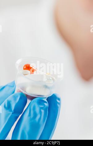 La mano del medico femminile in guanto blu che tiene i bicchieri graduati della medicina pieni di pillole. Copia spazio e sfondo bianco. Foto Stock