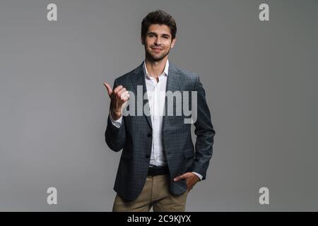 Foto di un uomo d'affari allegro in giacca sorridente e puntante dito da parte isolato su parete grigia Foto Stock