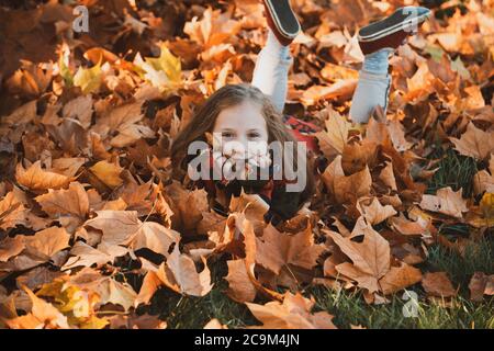 Carina bambina nel parco autunnale. Bambina in foglie. Felice bambino ridendo e giocando foglie in autunno all'aperto. Bella ragazza felice che si diverte dentro Foto Stock