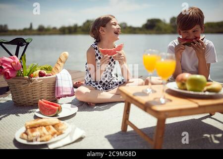 Fratello e sorella godono di mangiare l'anguria su un picnik sul molo del lago Foto Stock