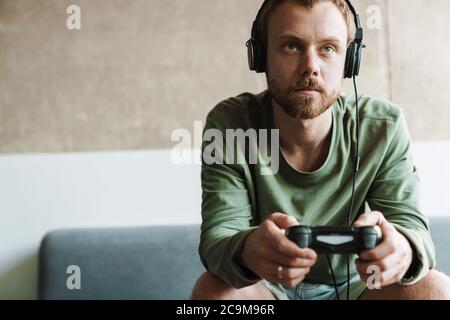 Foto di un uomo che indossa le cuffie che gioca a un videogioco con il joystick mentre sei seduto sul divano a casa Foto Stock