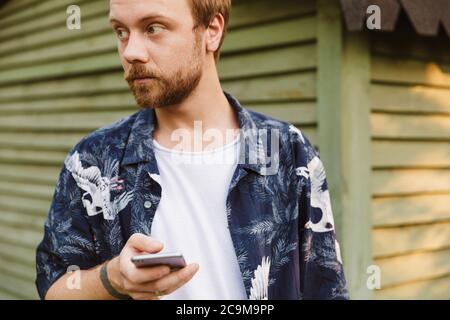 Immagine di un giovane uomo caucasico che tiene il cellulare mentre si trova in piedi da casa di legno all'aperto Foto Stock