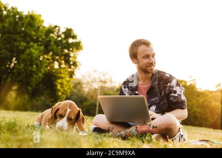 Immagine di un giovane uomo caucasico che usa un computer portatile mentre si siede con il suo cane beagle sull'erba nel parco estivo Foto Stock