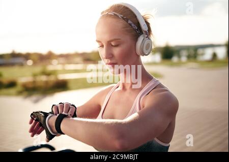 Fitness ragazza snella che indossa abiti sportivi utilizza un orologio intelligente e ascolta la musica con le sue cuffie, retroilluminata, parco soleggiato, città estiva Foto Stock