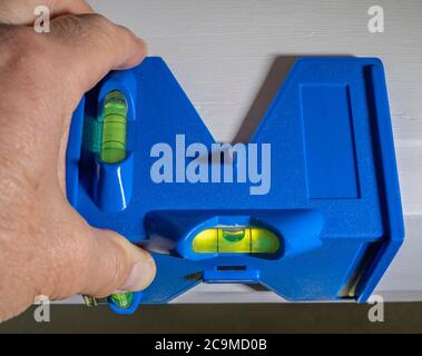 Chiudere il POV della mano di un uomo controllando la precisione orizzontale della cornice di una porta, con una livella a bolla / livella a bolla in plastica blu a forma di angolo. Foto Stock
