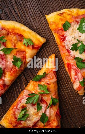 Fette di pizza calda con mozzarella, prosciutto, pomodoro e prezzemolo su tagliere di legno marrone, macro vista dall'alto Foto Stock